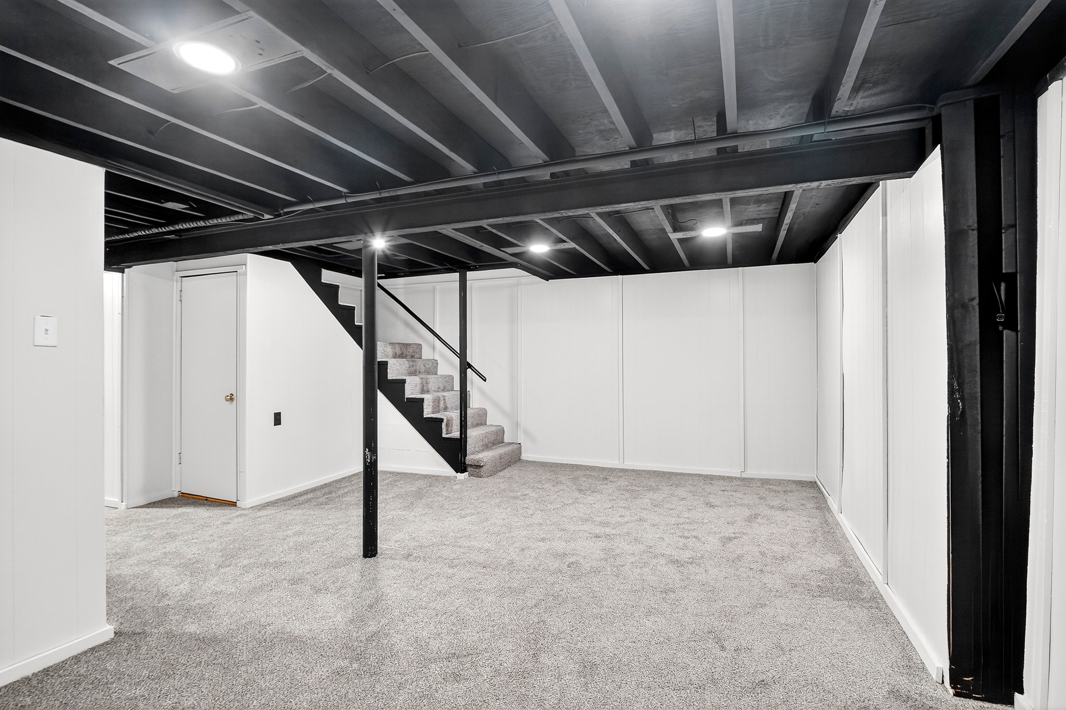 Basement with black ceiling beams, carpeted floor, and white walls.