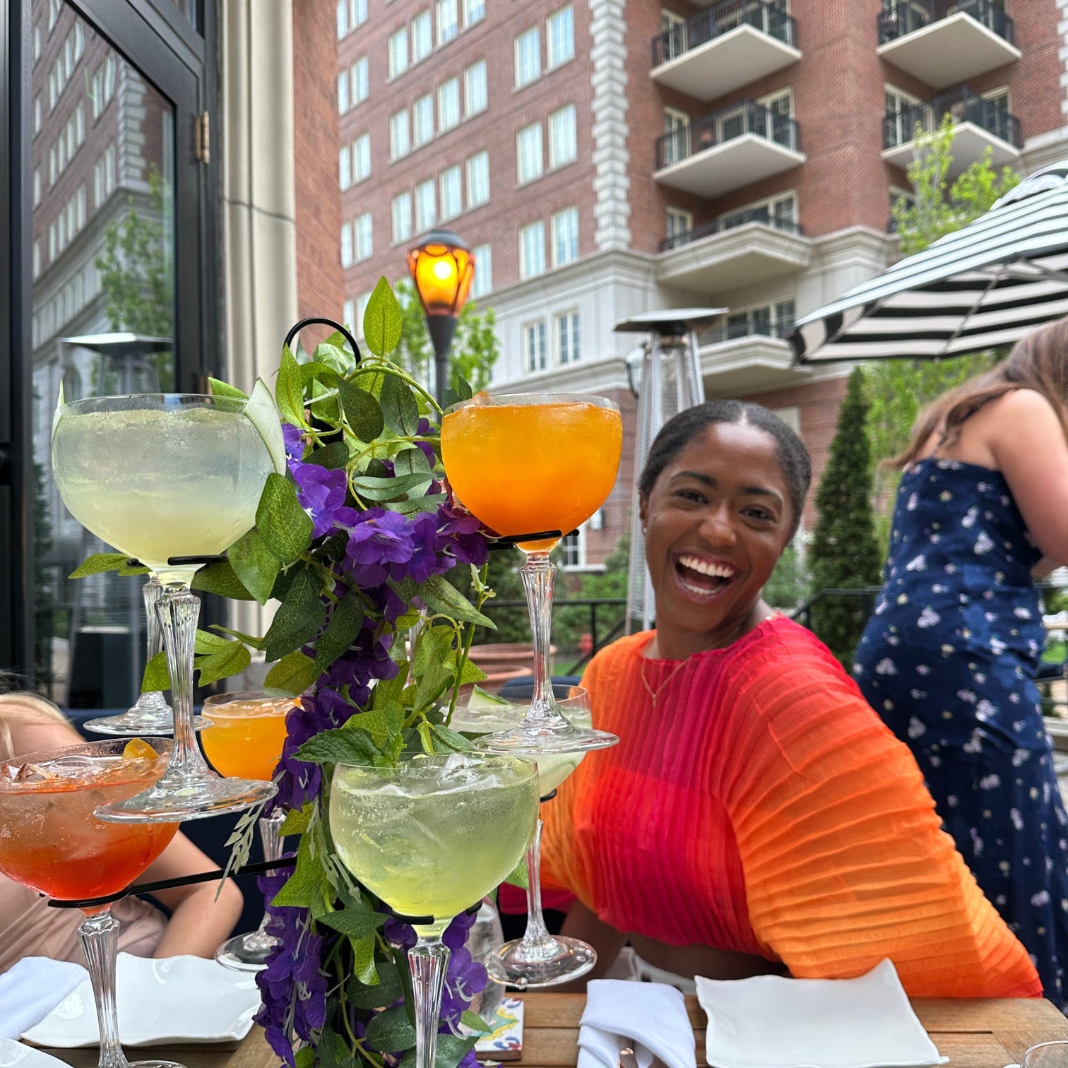 Woman sitting at an outdoor dining table with drinks and flowers, smiling.