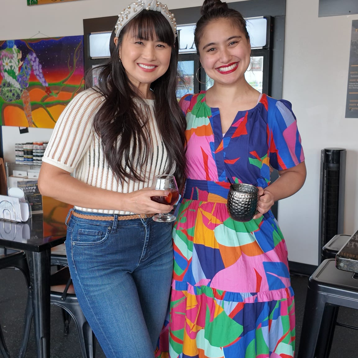 Two women standing together in a casual setting with a TV screen in the background.