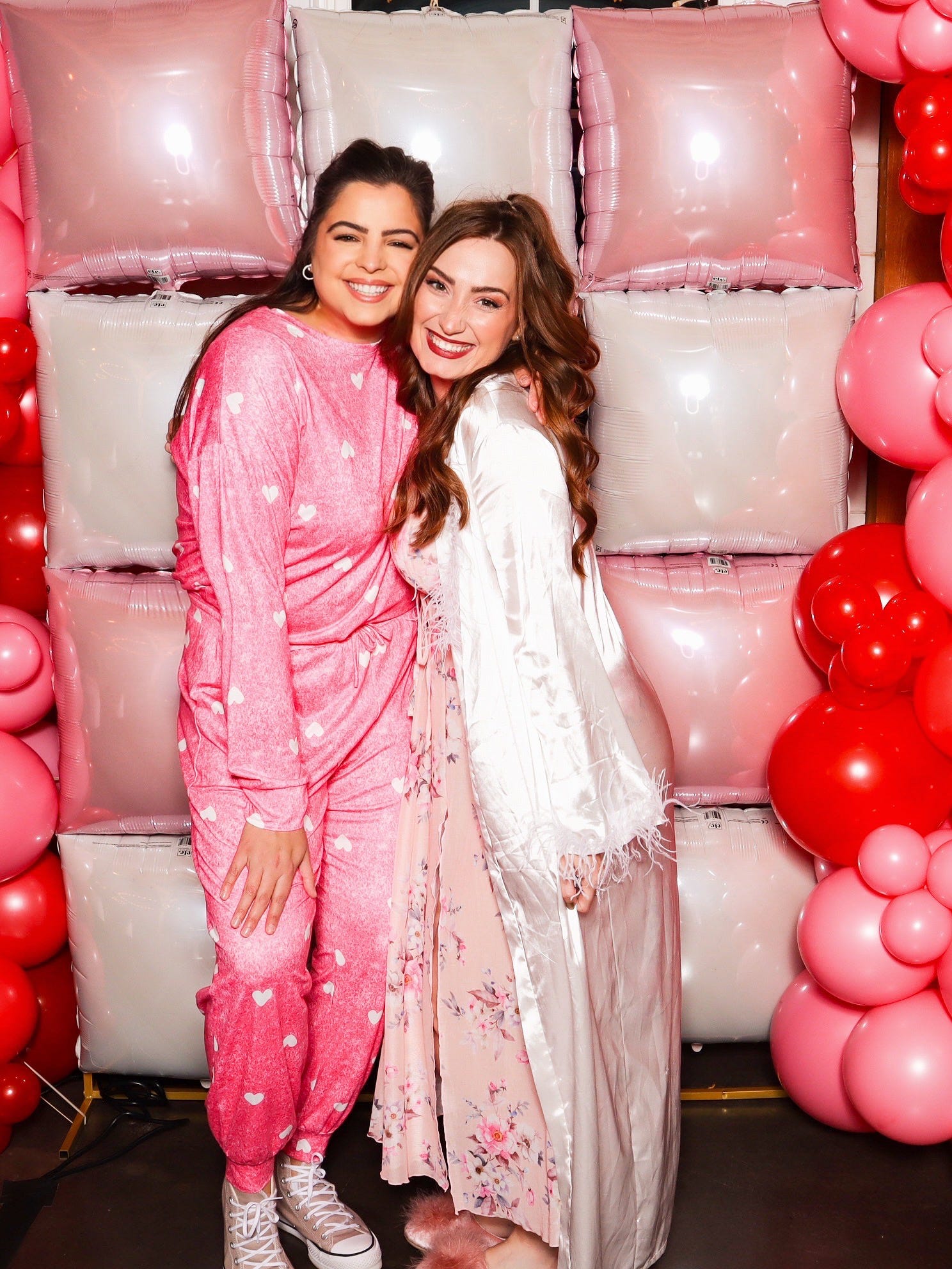 Two women posing together in front of pink and red balloons and cushions.