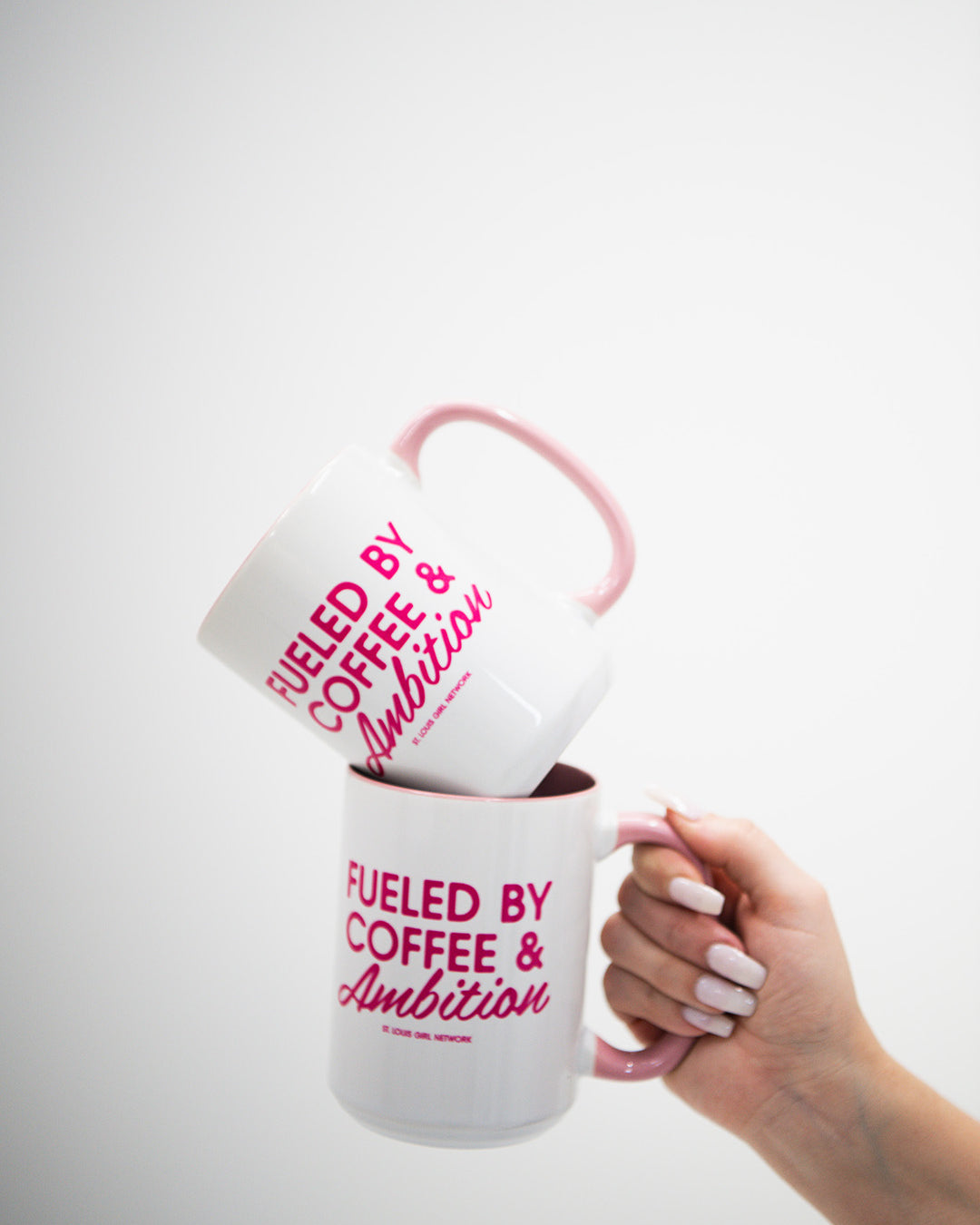Hand holding a white mug with pink text 'Fueled by Coffee & Ambition' against a white background