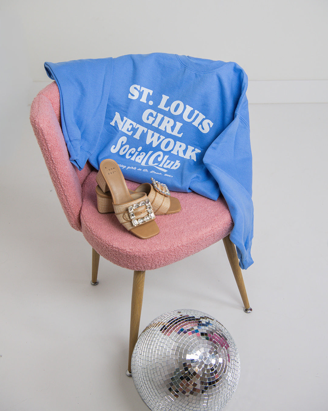Blue sweatshirt with text on a pink chair with shoes, a disco ball, and a white wall background
