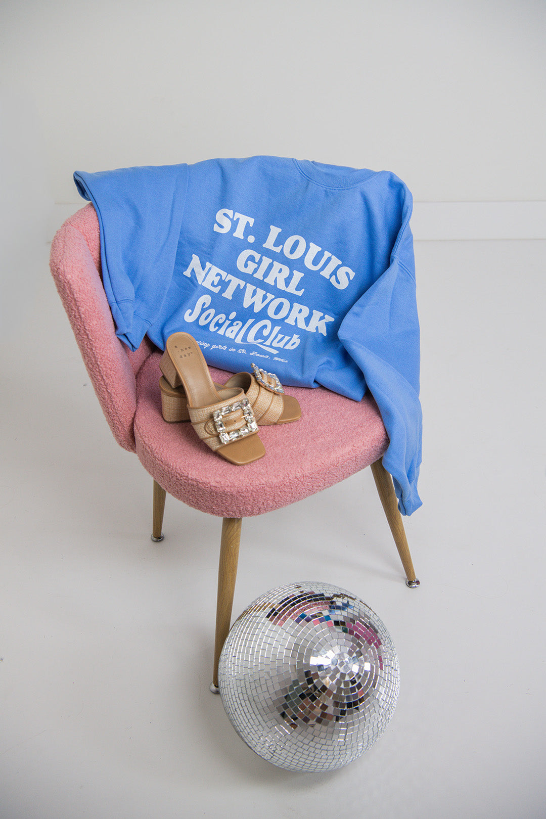 Blue sweatshirt with text on a pink chair with shoes, a disco ball, and a white wall background