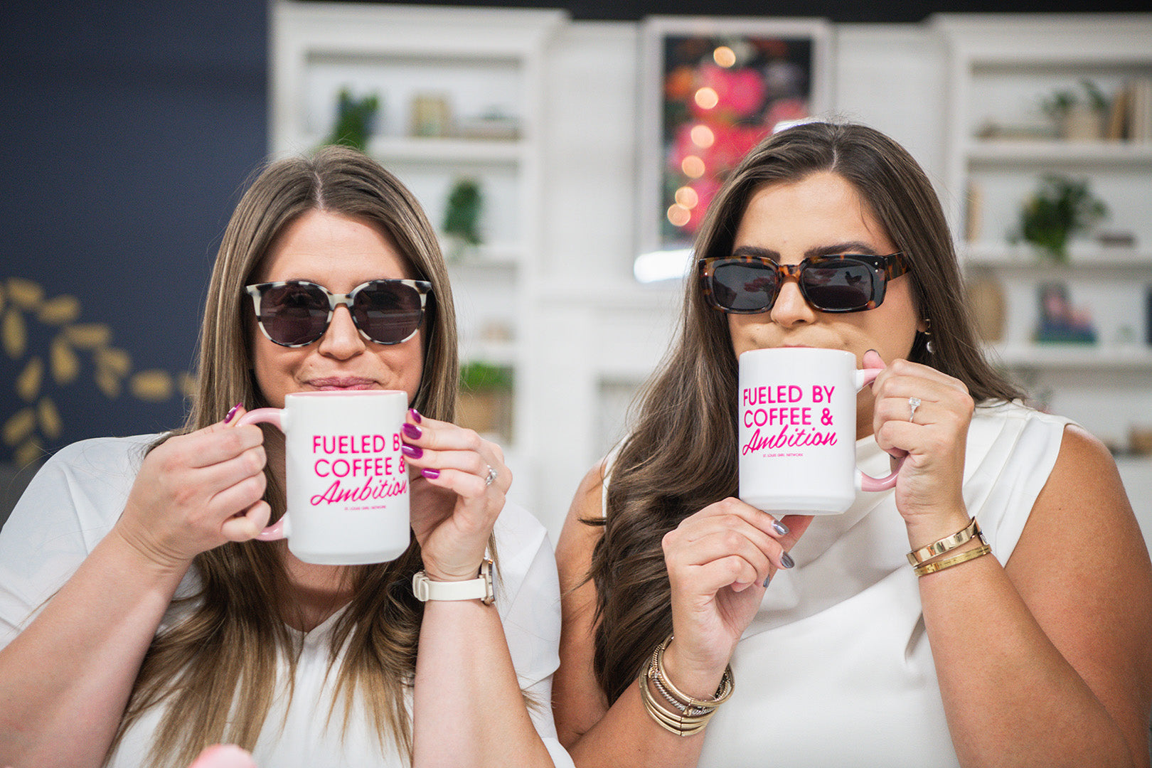 Two women holding mugs with 'Fueled by Coffee & Ambition' text in a casual setting.