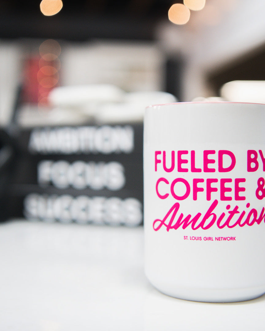 White mug with pink text 'Fueled by Coffee & Ambition' on a blurred background