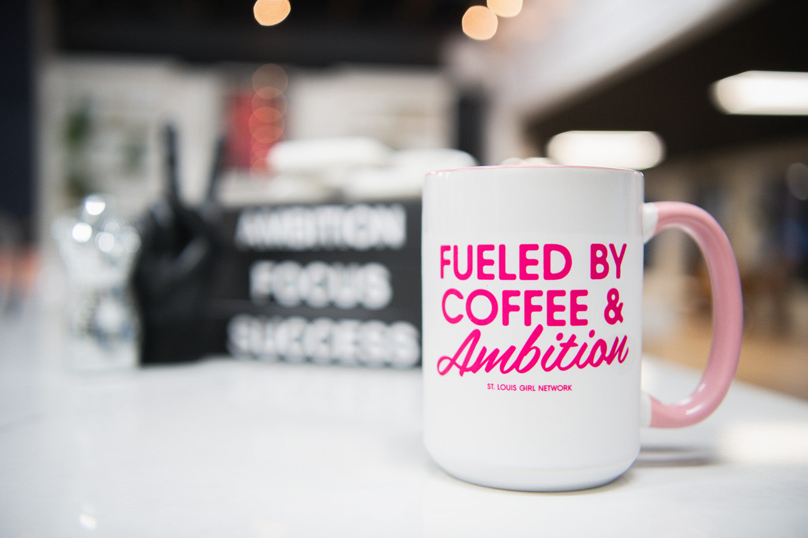 White mug with pink text 'Fueled by Coffee & Ambition' on a blurred background