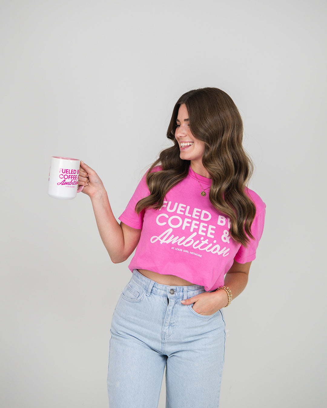 Woman wearing a pink t-shirt with text "Fueled by Coffee & Ambition", holding a white mug with text, on a plain background