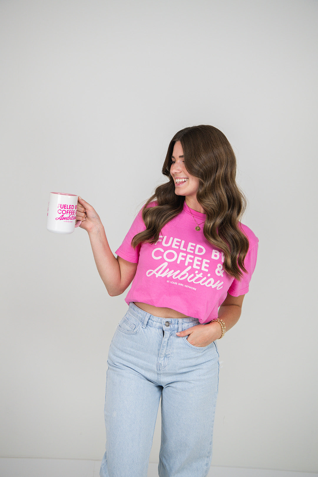 Woman wearing a pink t-shirt with text "Fueled by Coffee & Ambition", holding a white mug with text, on a plain background