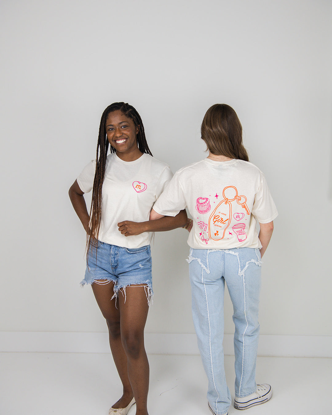 Two women wearing beige t-shirts with colorful graphics, one facing forward and the other facing backward, on a plain background.
