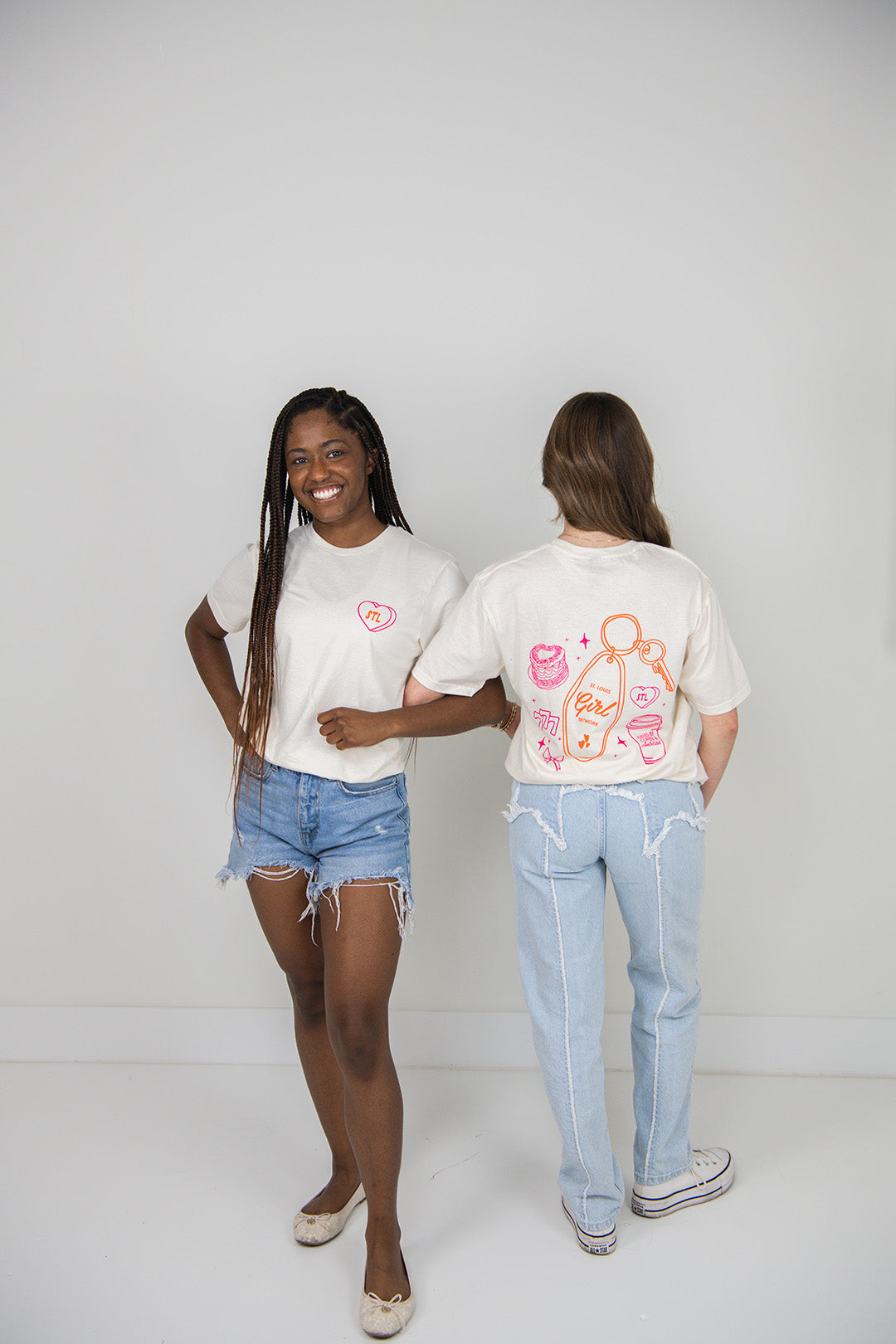 Two women wearing beige t-shirts with colorful graphics, one facing forward and the other facing backward, on a plain background.
