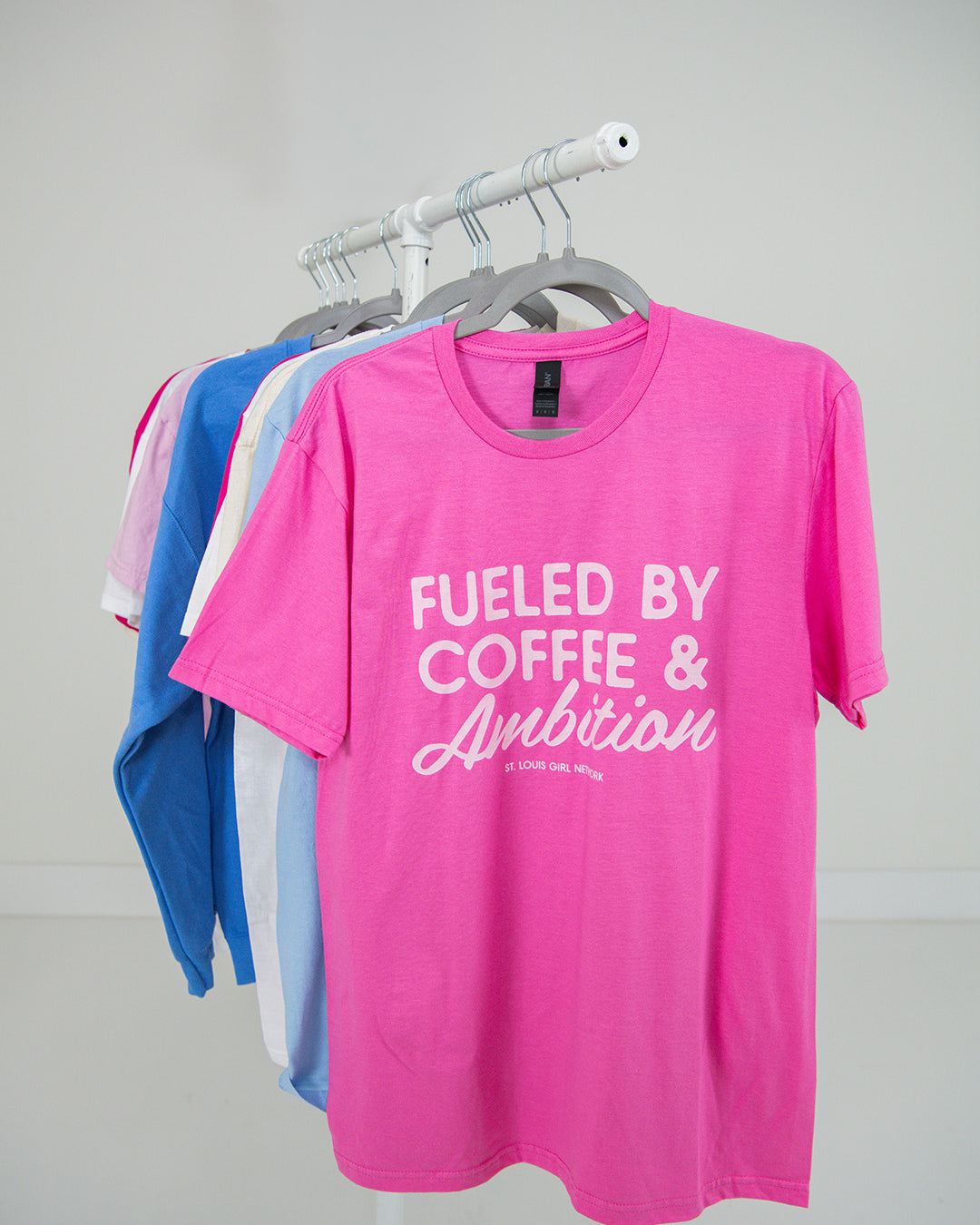 Pink t-shirt with 'Fueled by Coffee & Ambition' text on a rack against a plain background