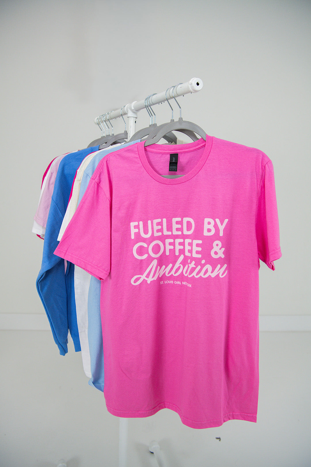 Pink t-shirt with 'Fueled by Coffee & Ambition' text on a rack against a plain background