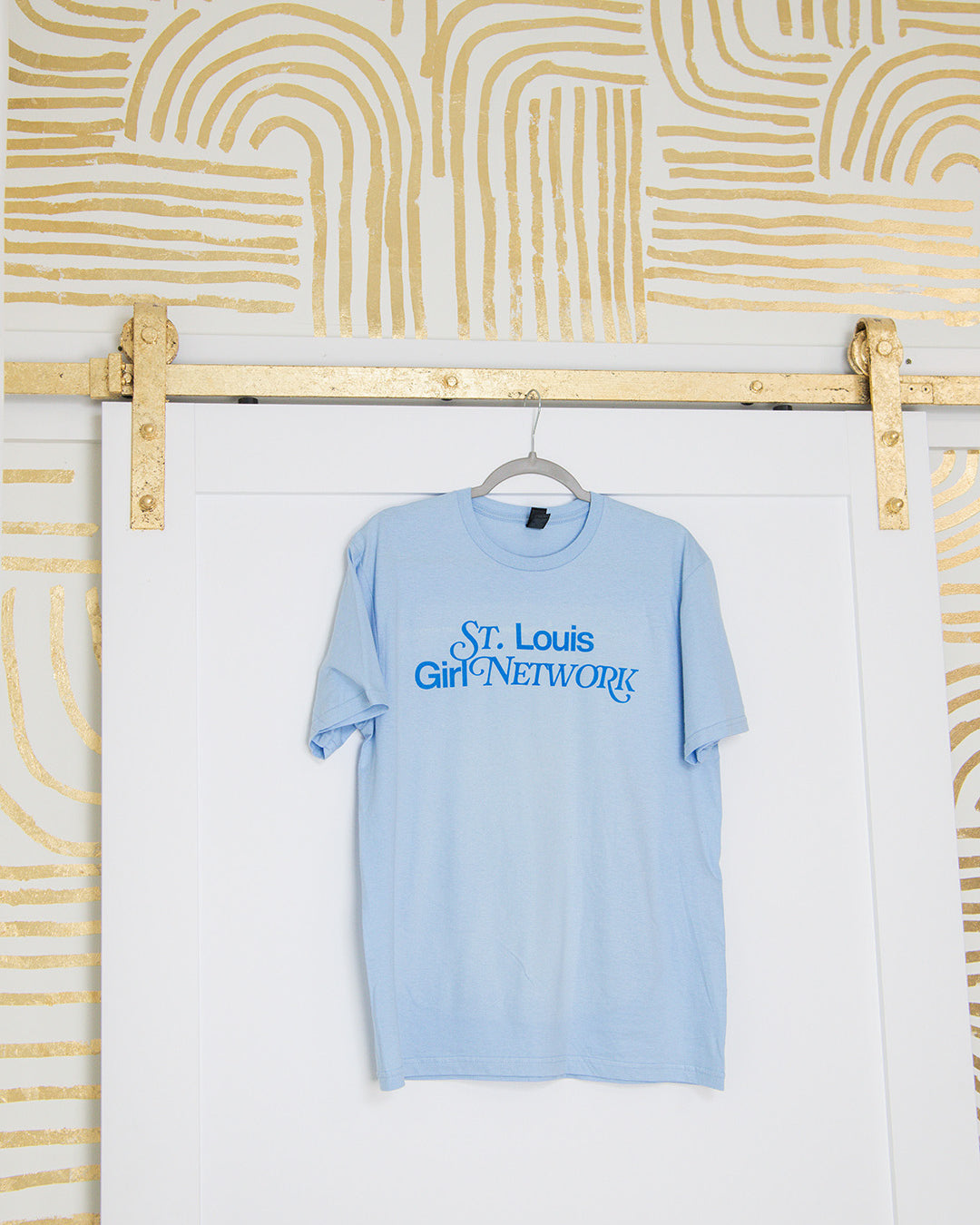 Light blue t-shirt with 'St. Louis Girl Network' text hanging on a rack against a white wall with gold abstract patterns.