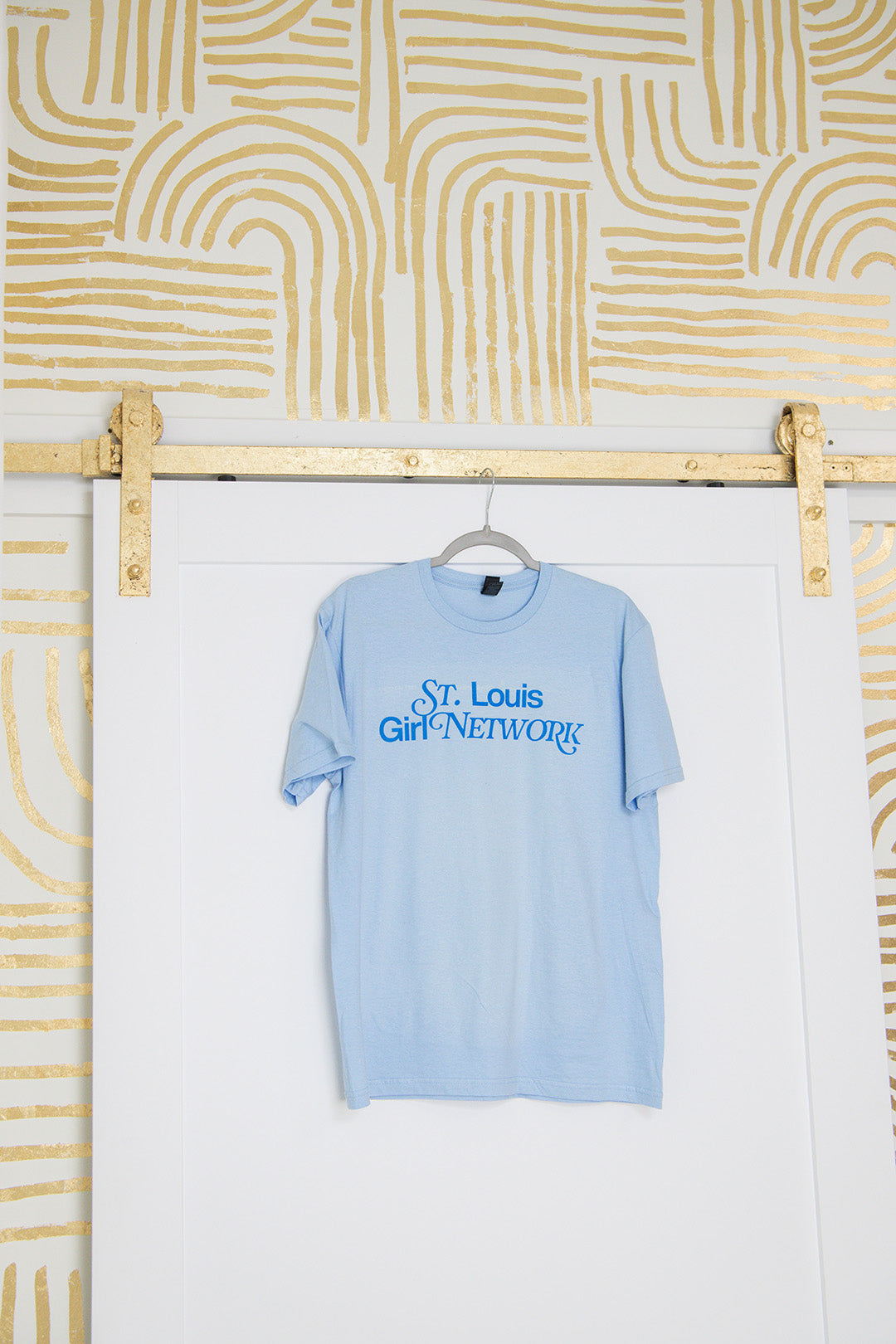 Light blue t-shirt with 'St. Louis Girl Network' text hanging on a rack against a white wall with gold abstract patterns.