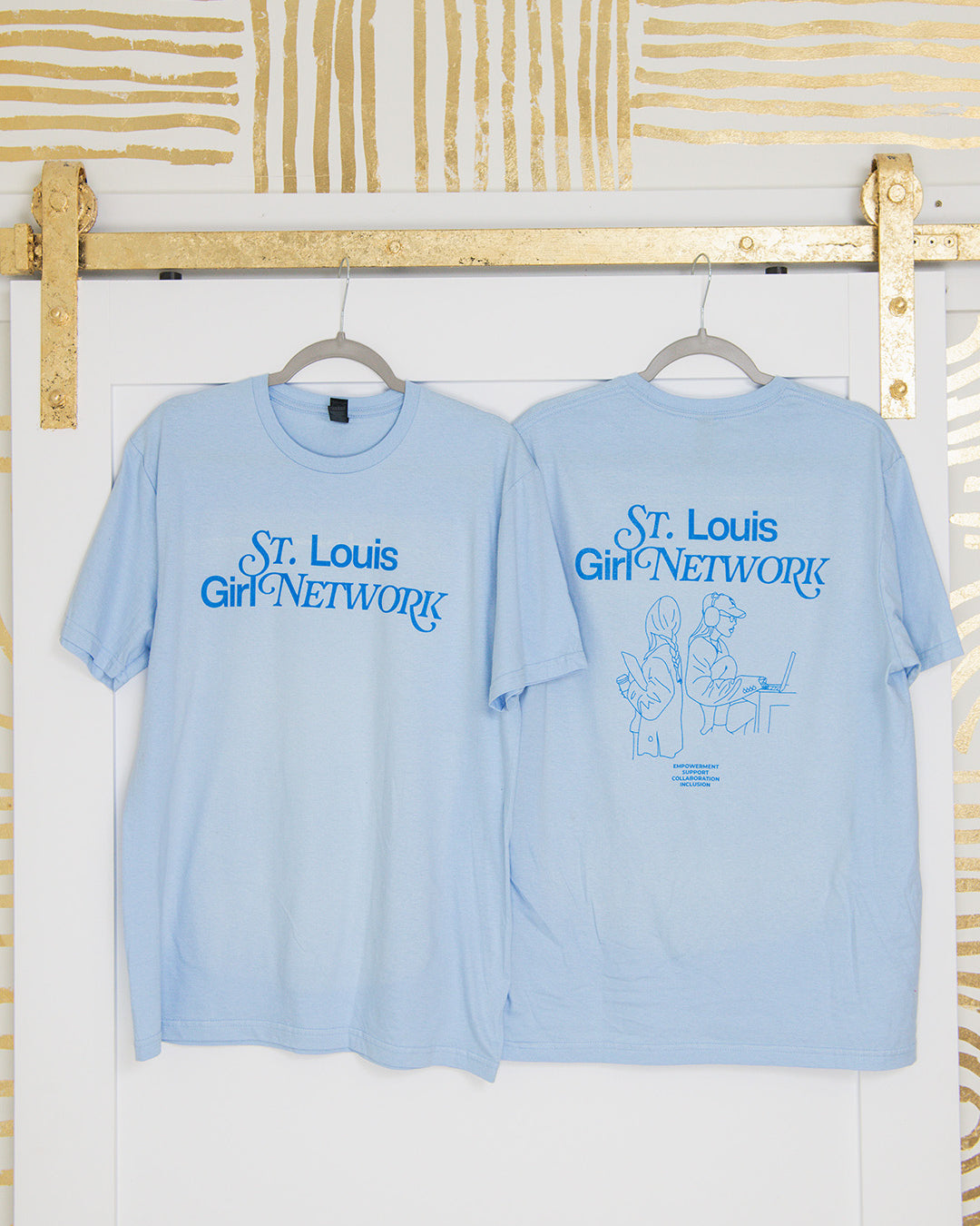 Two light blue t-shirts with 'St. Louis Girl Network' text on hangers against a white wall with gold abstract patterns.