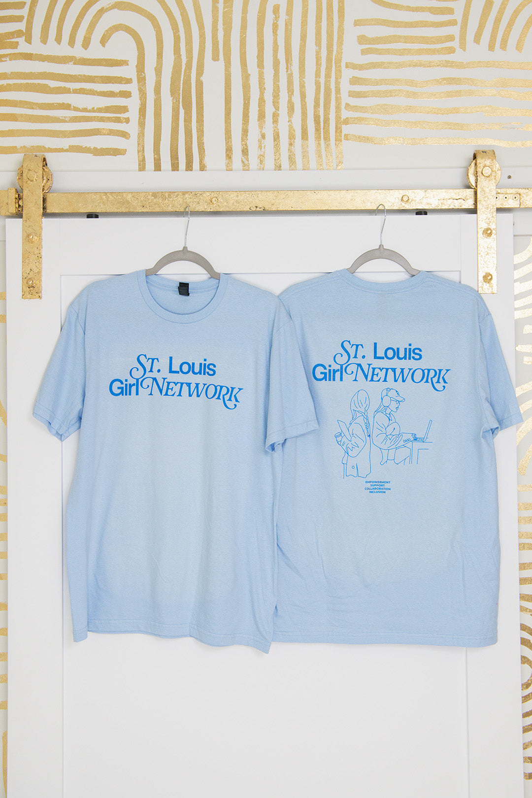 Two light blue t-shirts with 'St. Louis Girl Network' text on hangers against a white wall with gold abstract patterns.