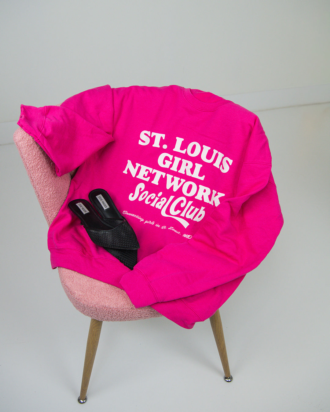 Pink sweatshirt with 'St. Louis Girl Network Social Club' text on a chair against a white background