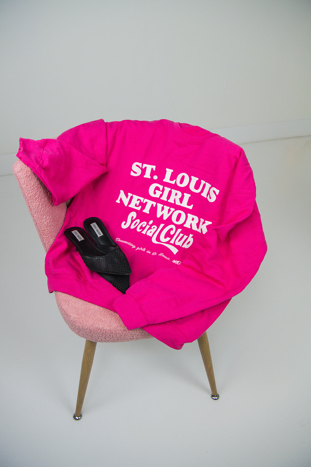Pink sweatshirt with 'St. Louis Girl Network Social Club' text on a chair against a white background