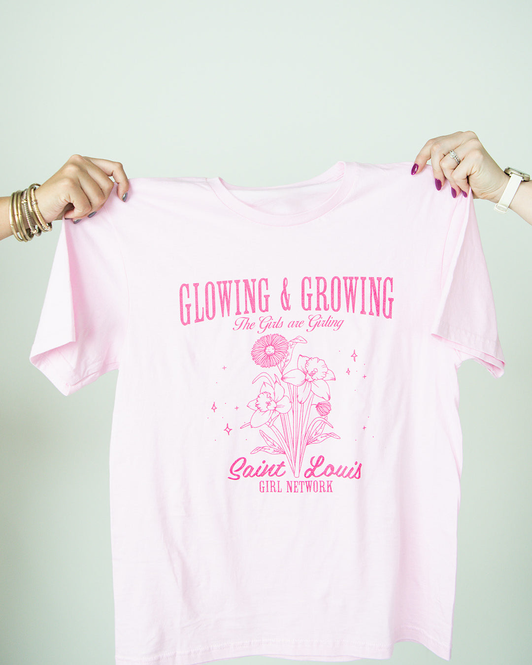 Pink t-shirt with text 'Glowing & Growing' and graphics held by two hands against a white background