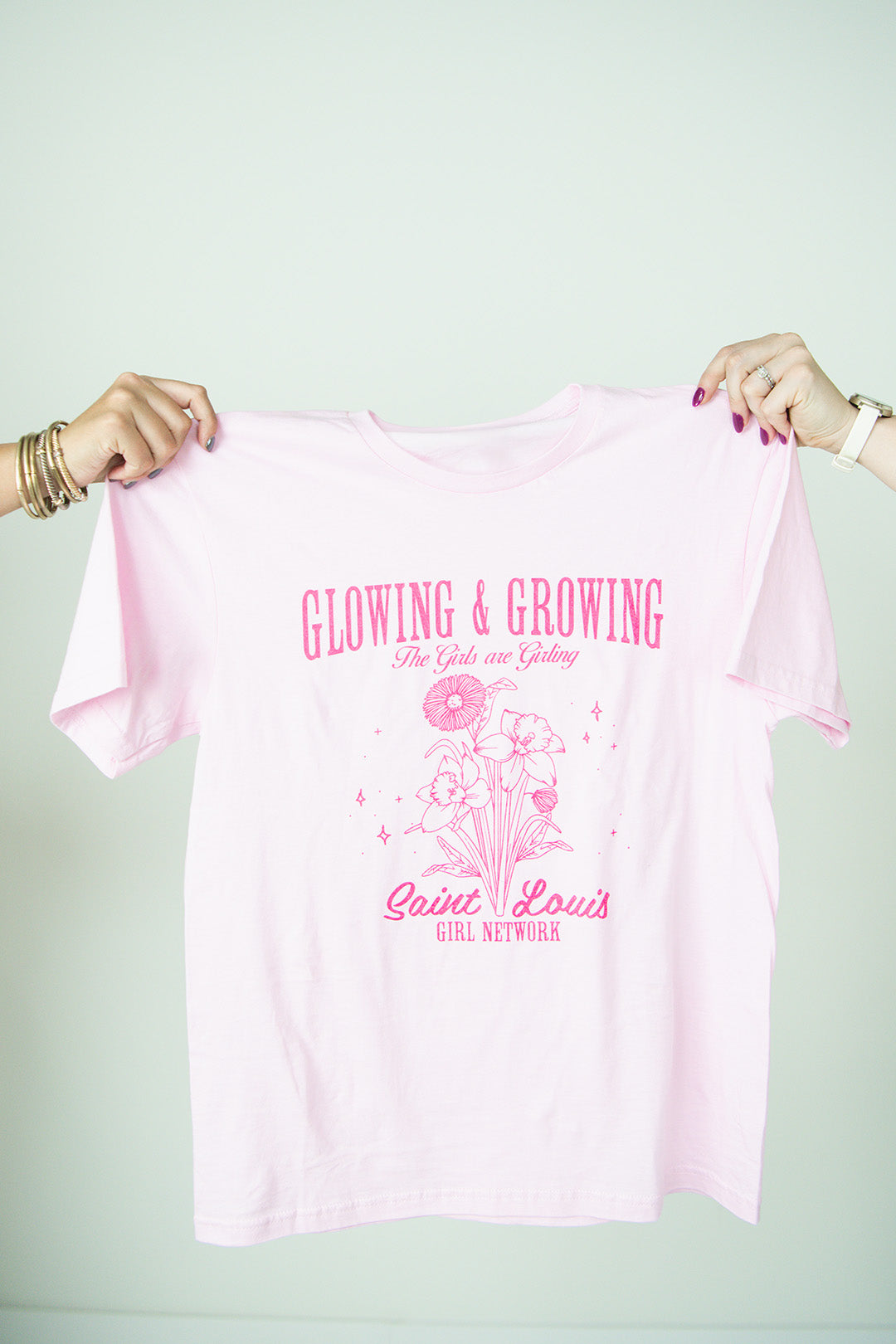 Pink t-shirt with text 'Glowing & Growing' and graphics held by two hands against a white background
