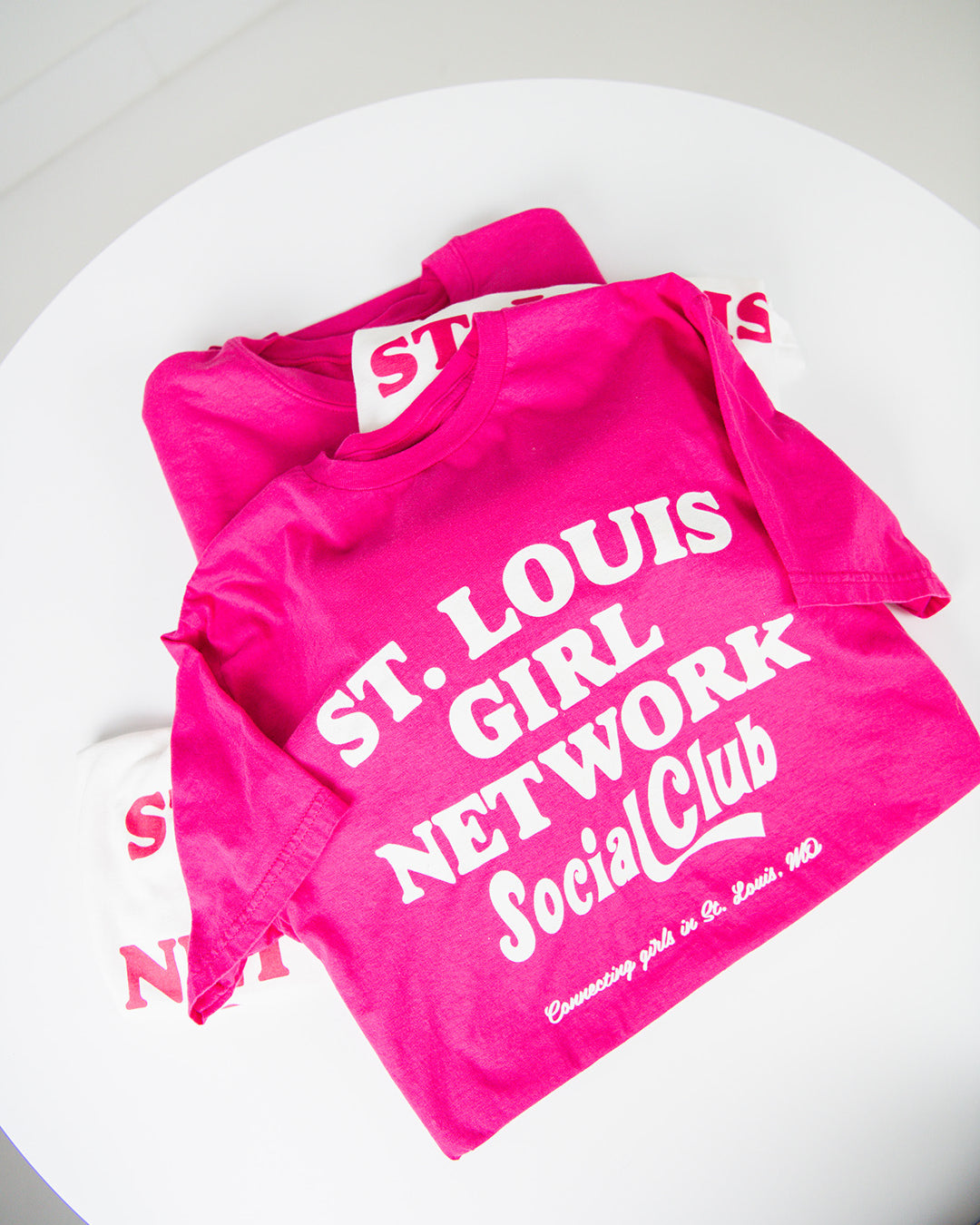 Pink t-shirt with 'St. Louis Girl Network Social Club' text on a white surface