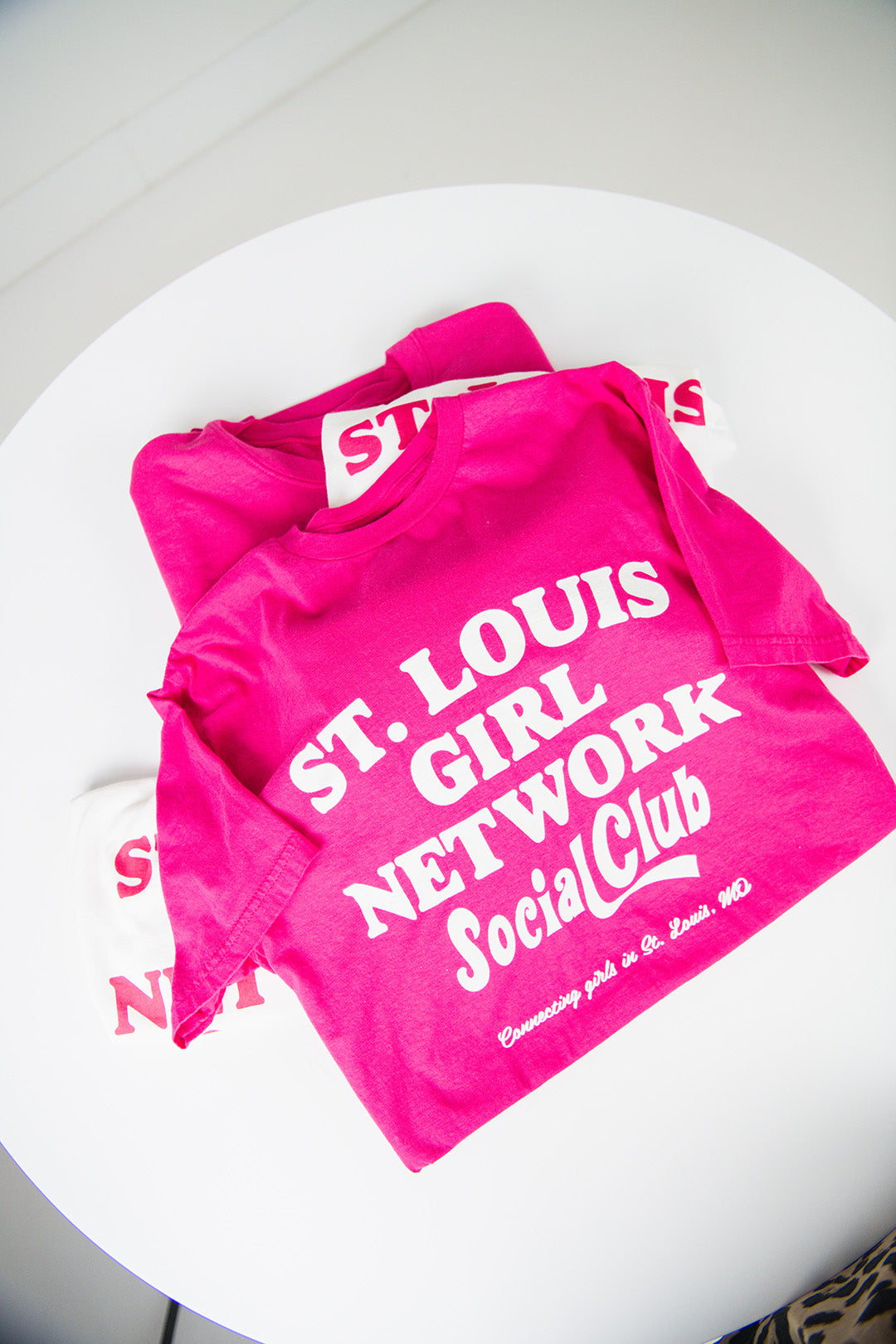 Pink t-shirt with 'St. Louis Girl Network Social Club' text on a white surface