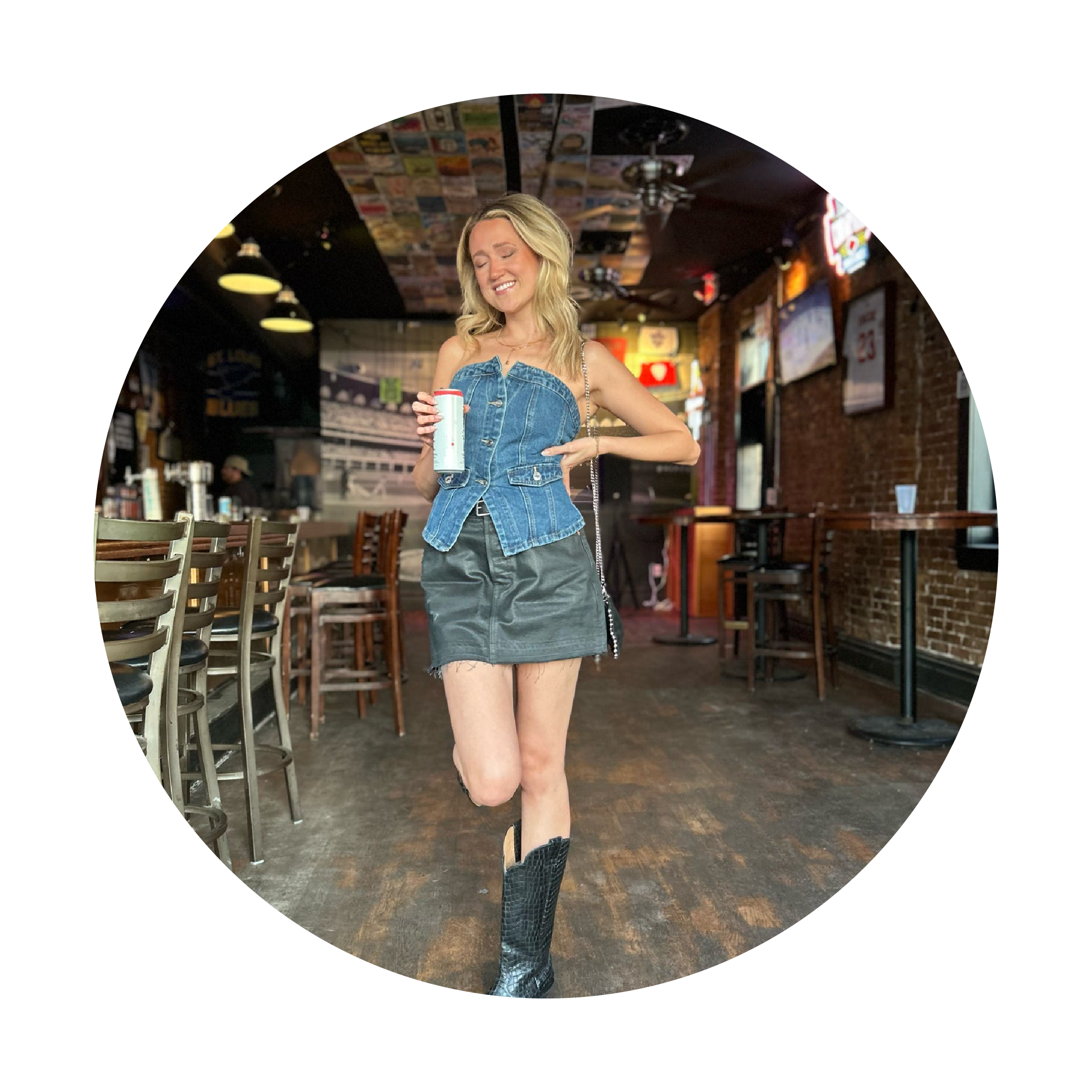Woman in a bar wearing a denim top and skirt with cowboy boots, holding a drink.