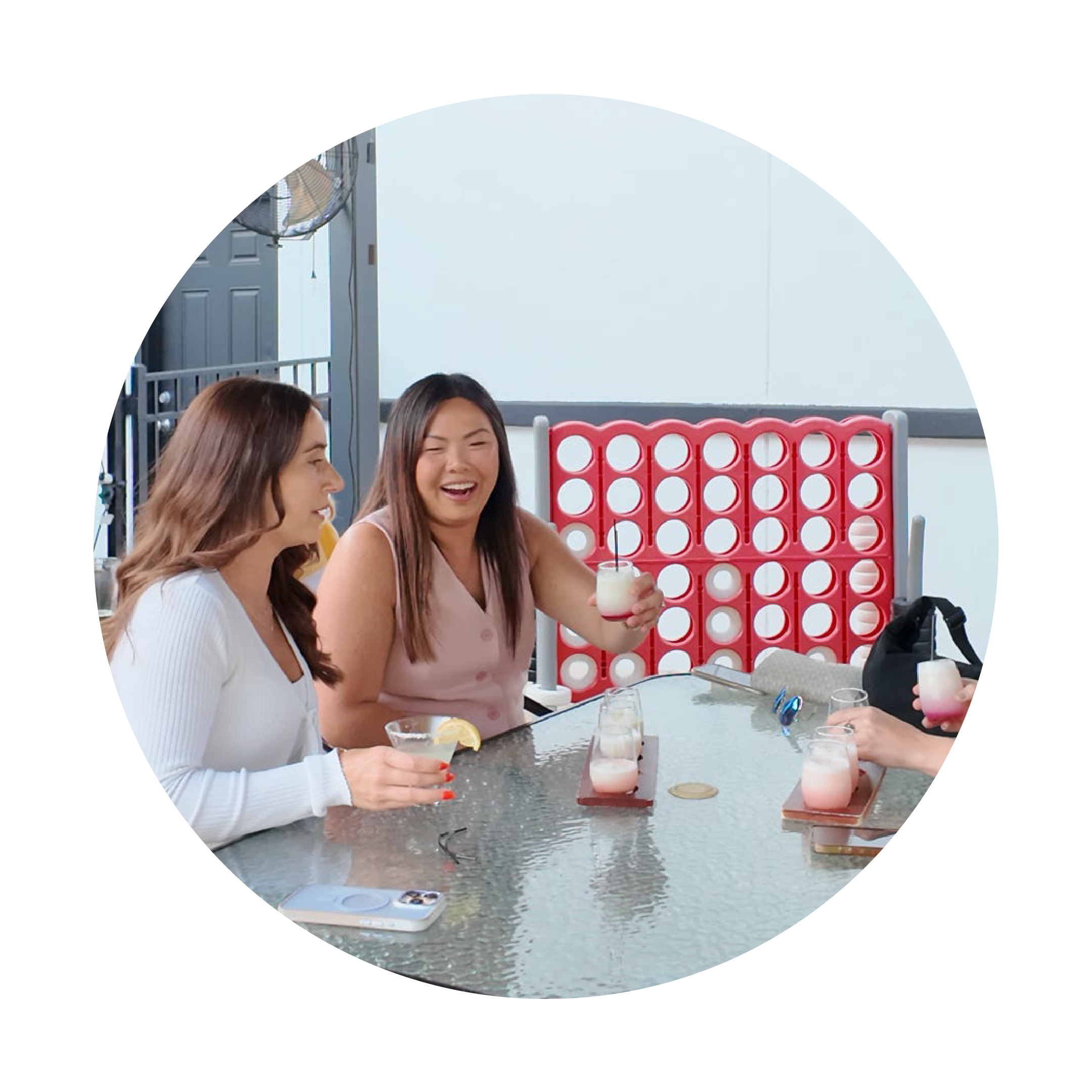 Two women sitting at a table with drinks, smiling and laughing, in front of a red and white game.