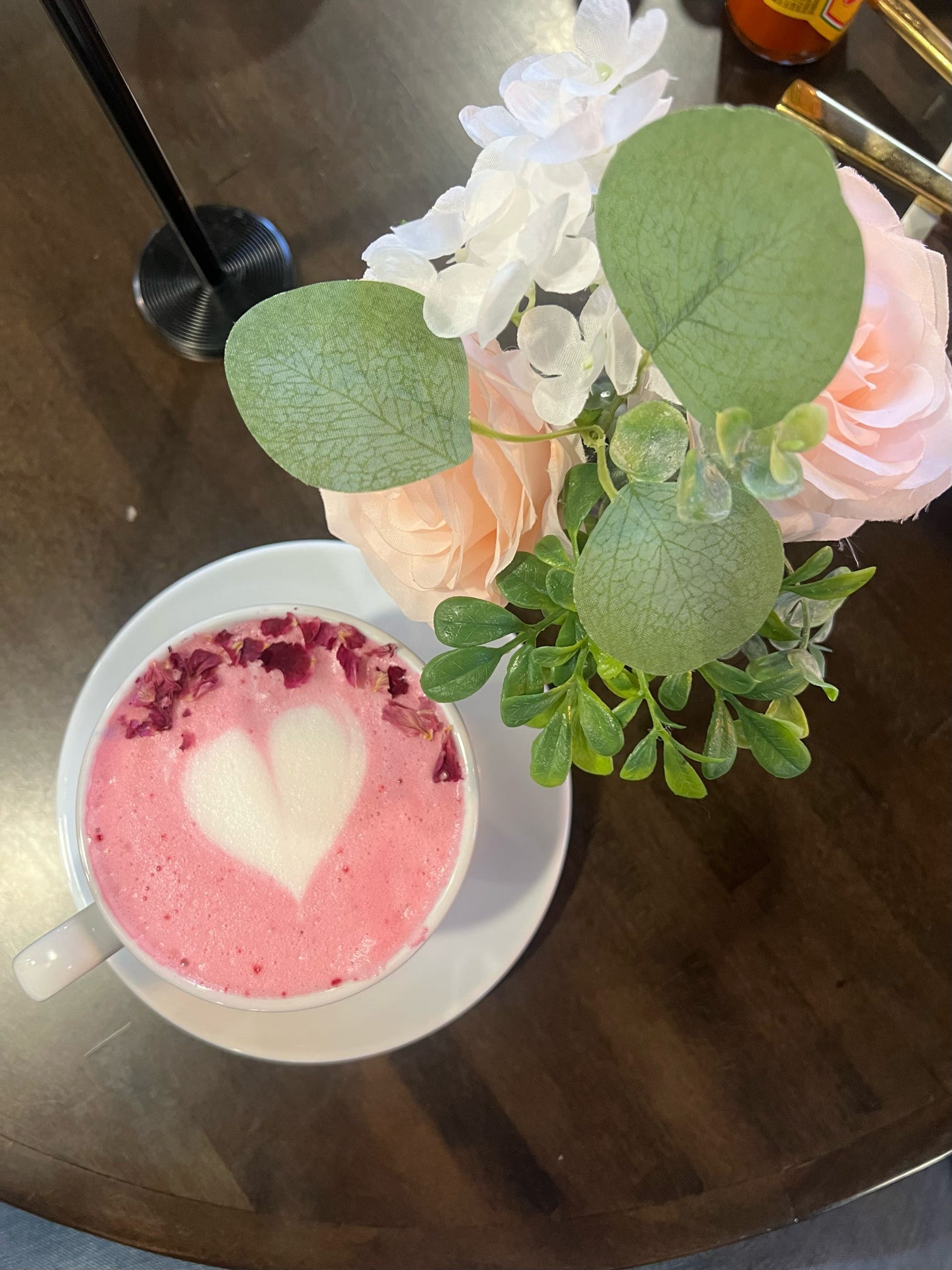 Pink drink with a heart foam in a white cup, accompanied by a small plant and flowers on a reflective surface.