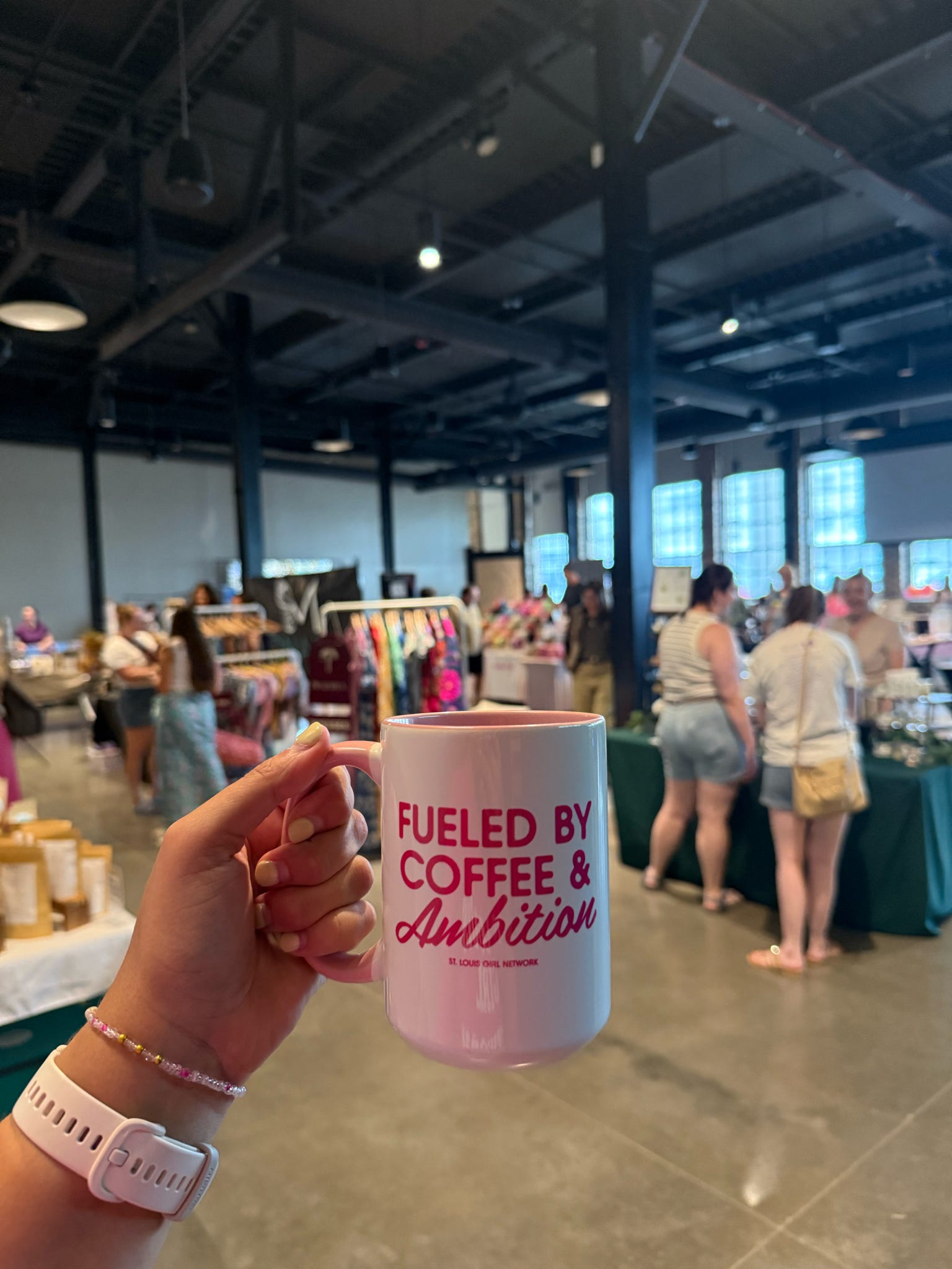 Person holding a pink mug with 'Fueled by Coffee & Ambition' text in an indoor setting with people and tables.
