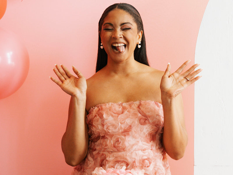 Woman in a pink dress standing next to a cake with pink balloons in the background