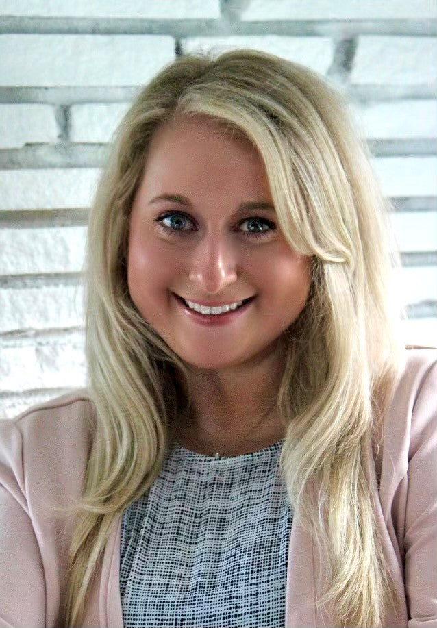 Woman with blonde hair and a pink jacket smiling in front of a white brick wall.