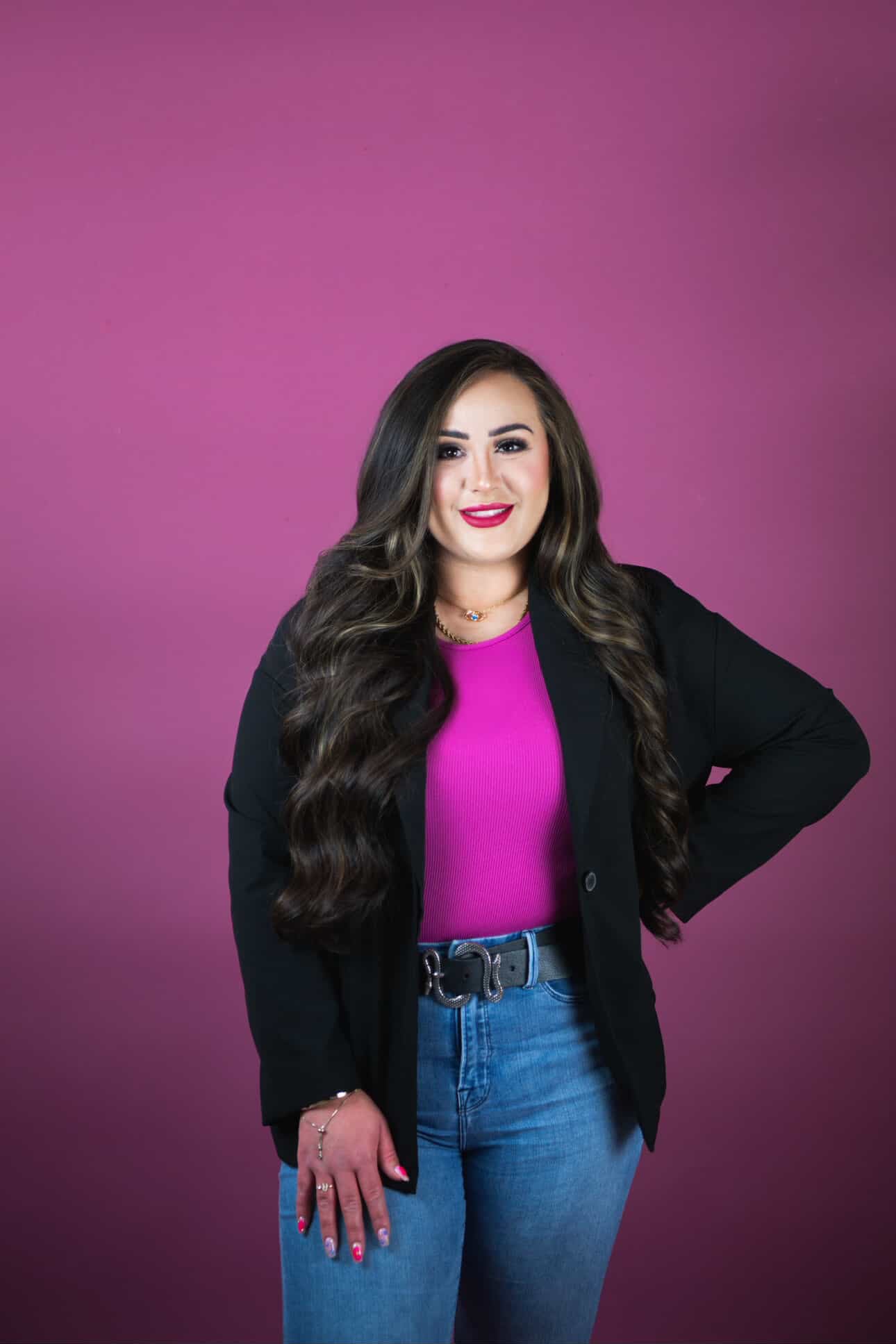 Woman wearing a black blazer, pink top, and blue jeans against a purple background