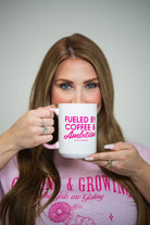 Woman holding a mug with 'Fueled by Coffee & Ambition' text, wearing a pink shirt.