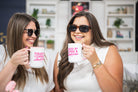 Two women holding mugs with text, smiling in a casual setting.