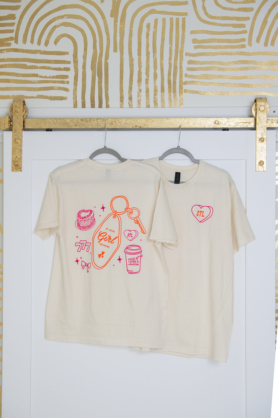 Beige t-shirt with graphic designs on a white wall with gold abstract patterns